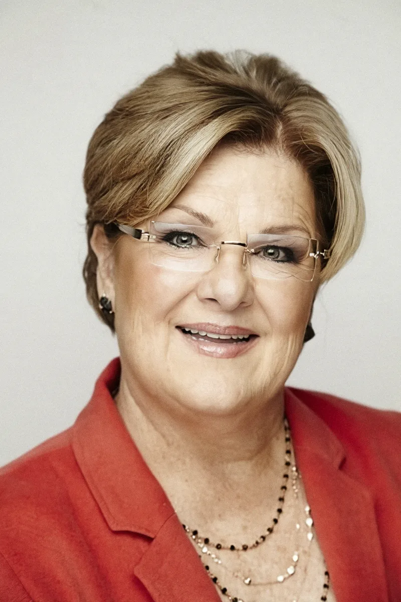 Close portrait of a smiling woman with blonde hair, glasses and a red jacket against a white background.