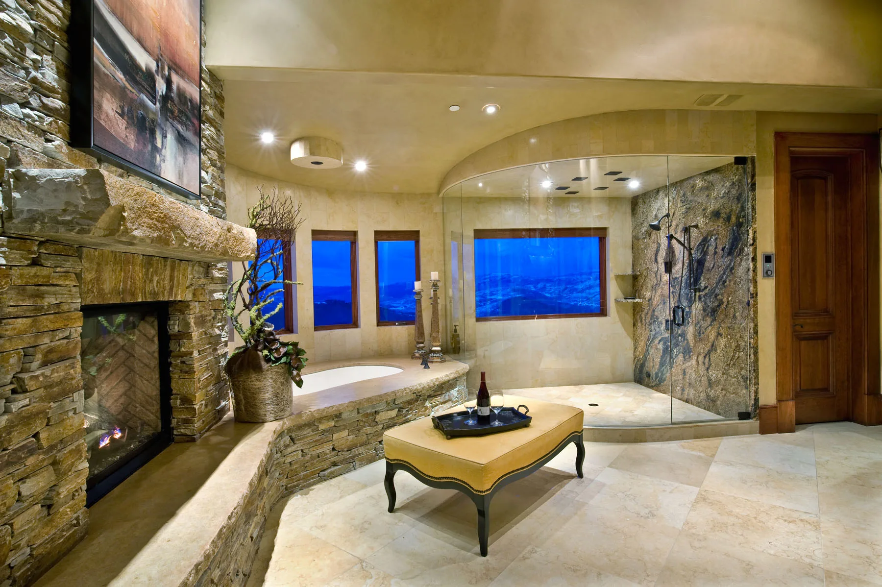 Bathroom with an oval soaking tub, curved stone walls, a fireplace and windows showing a blue evening sky