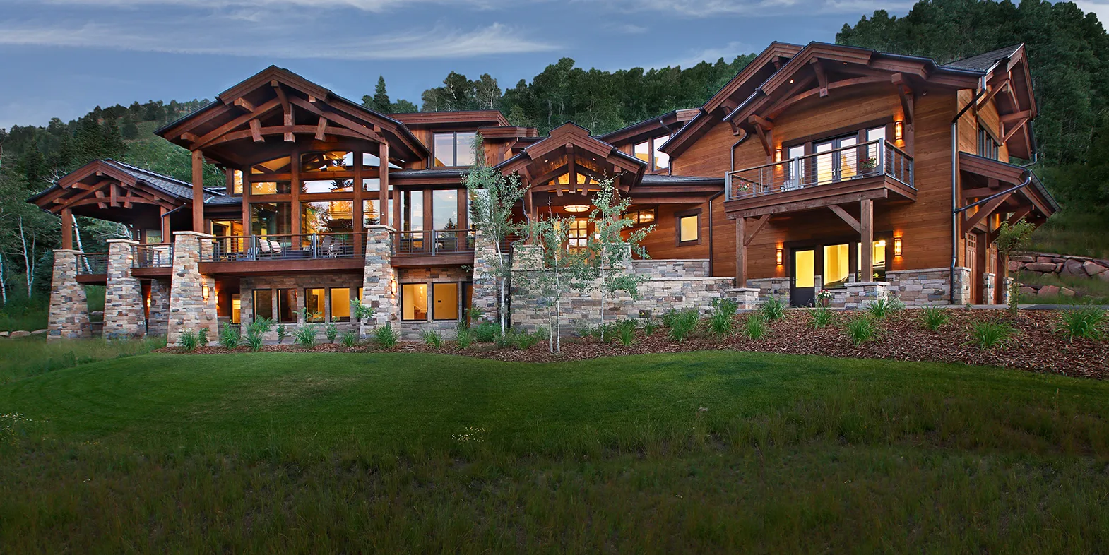 Dusk exterior of a two-story stone and timber mountain home with illuminated windows and a snow-capped peak behind