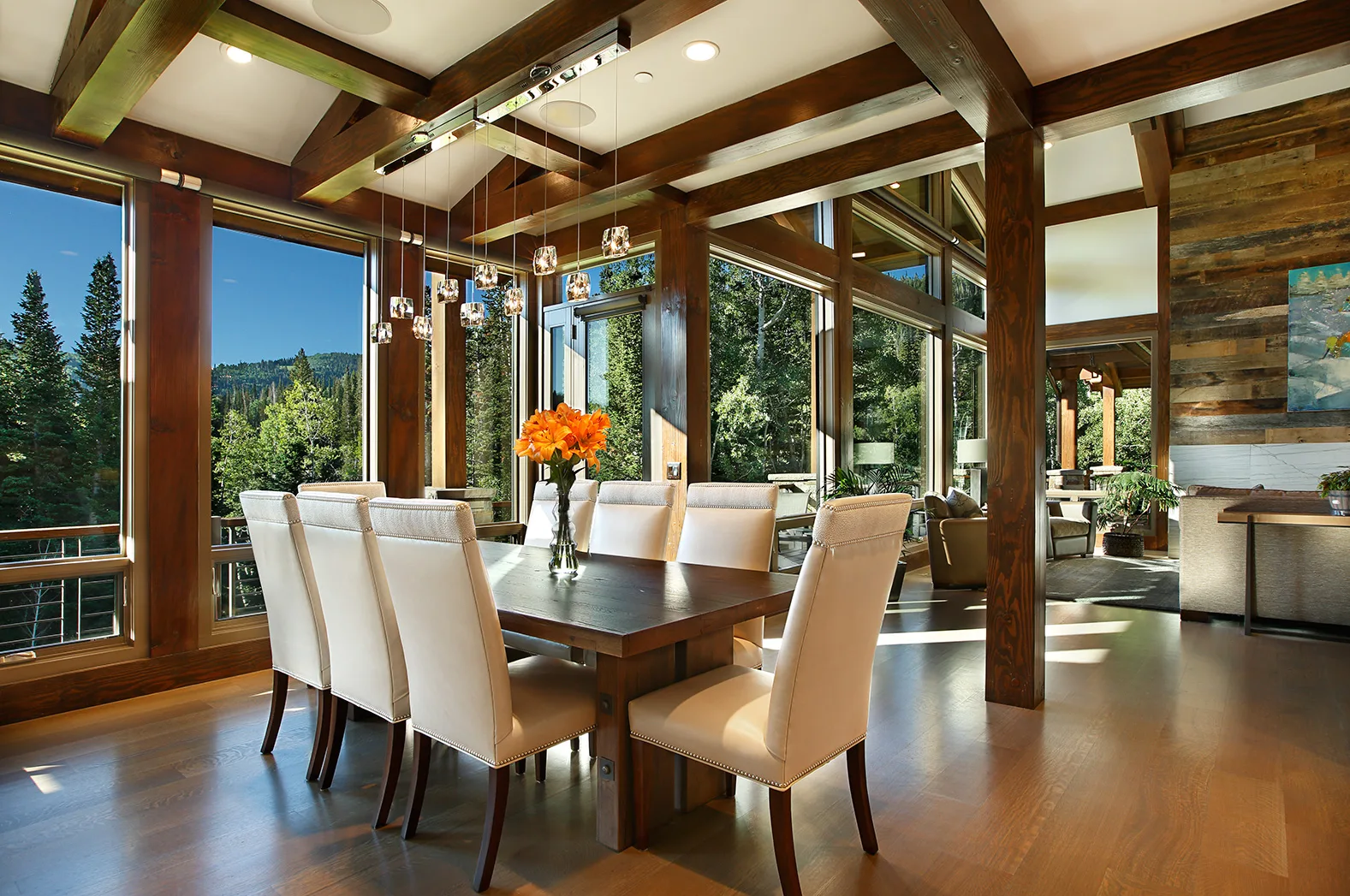 Dining room with a long table, white upholstered chairs and fresh flowers beneath floor-to-ceiling glass and timber trusses facing the forest